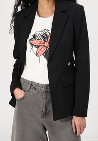 Femme portant un blazer noir sur un t-shirt blanc avec des cheveux noirs et un motif de fleur rouge, associé à un jean denim gris taille haute.