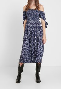 Marineblå blommig midiklänning med off-shoulder ärmar, smockad bodice och knytdetaljer. Kombinerad med svarta cowboystövlar för kontrast.
