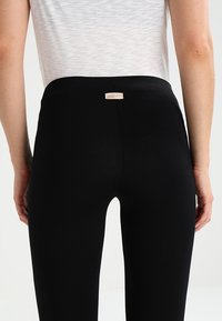 Leggings noirs en tissu extensible, avec une taille mi-haute et une petite étiquette logo beige à l'arrière. Texture lisse, design près du corps.