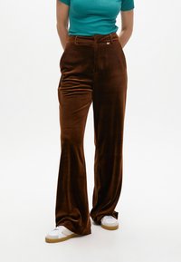 Pantaloni marrone in velluto a vita alta con gamba larga, caratterizzati da una chiusura a bottone frontale e una texture morbida, abbinati a sneakers bianche.