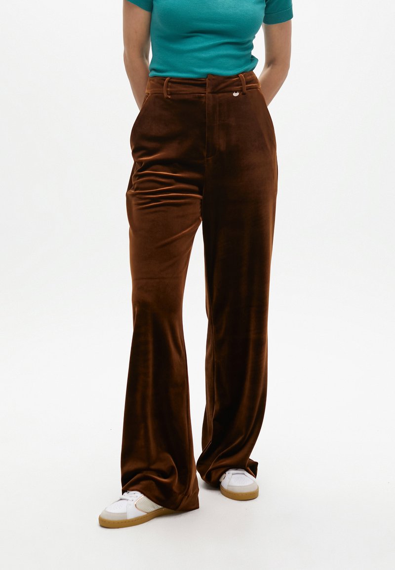 Pantaloni marrone in velluto a vita alta con gamba larga, caratterizzati da una chiusura a bottone frontale e una texture morbida, abbinati a sneakers bianche.