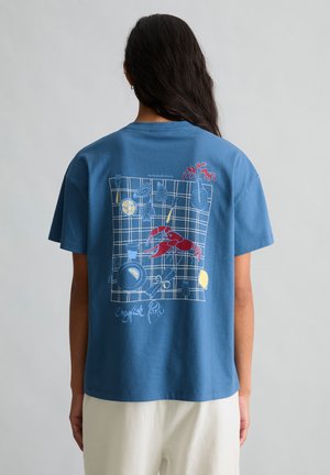 Persoon draagt een blauw overhemd met afbeelding van rode rivierkreeft, schijfjes citroen, oesters, bestek en vaas op geruit tafelkleed.