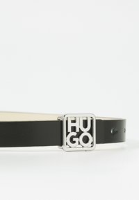 Musta nahkavyö, jossa on hopeinen neliönmuotoinen solki, jossa on brändin nimi "HUGO" lihavoidulla tekstillä. Sujuva pinta, minimalistinen muotoilu.