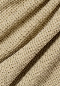 Beige geripptes Gewebe mit einem strukturierten, gewebten Muster, das eine weiche, flexible Qualität aufweist. Die Stoffstücke überlappen sich und zeigen Tiefe und Kontur.