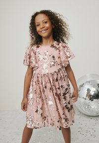 Lyserød kjole med korte, flæseærmer. Dækket af palietter og blomster mønstre. Tylleskørtet tilføjer volumen. Beliggende nær en discokugle.