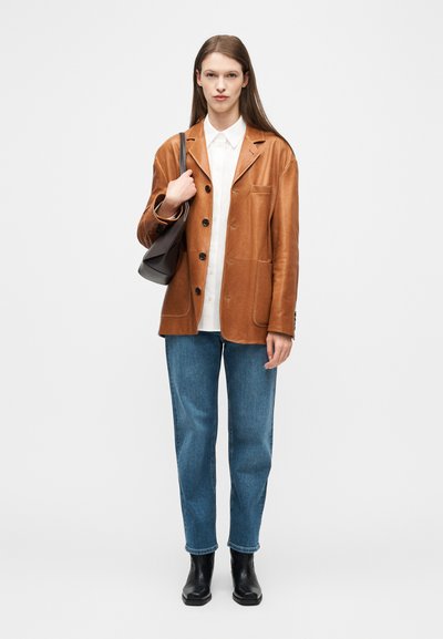 Femme portant une veste en cuir marron sur une chemise blanche, un jean bleu, des bottes noires, et portant un sac à bandoulière noir, debout devant un fond uni.