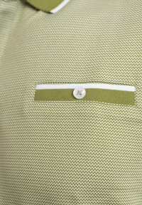 Polo de color verde con patrón que presenta un bolsillo con una franja blanca y un botón blanco. La textura es tejida con un diseño fino y repetitivo.