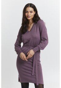 Abito a maniche lunghe con scollo a V in morbido maglione viola; presenta una cintura annodata in vita e spacchi laterali per facilitare i movimenti.