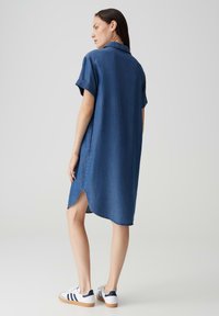 Denimhemdkleid in Dunkelblau, mit kurzen Ärmeln, knöpfbarem Kragen und seitlichen Schlitzen. Kombiniert mit weißenSneakern mit marineblauen Streifen.