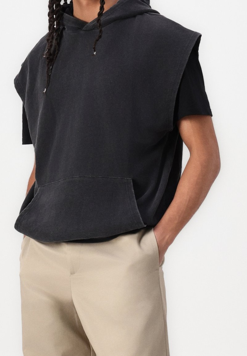 Personne portant un sweat à capuche noir sans manches par-dessus un t-shirt noir à manches courtes et un pantalon beige, les mains dans les poches.