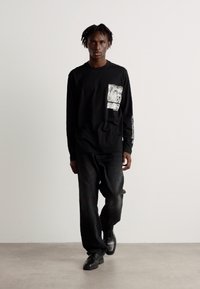 Zwart langoe mouwen shirt met een patroon op de zak, gecombineerd met donkere denim broek en zwarte schoenen. Eenvoudig ontwerp op een effen achtergrond.
