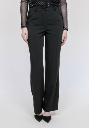 Pantalon noir à taille haute et jambes larges porté avec des chaussures noires à bouts pointus et un haut transparent noir à manches longues avec des pois blancs.