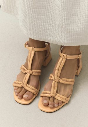 Bare feet wearing beige braided leather strappy sandals with block heels, paired with a white textured skirt.