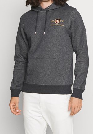 Mężczyzna ubrany w ciemnoszarą bluzę z kapturem z przednią kieszenią i logo "GANT American Sportswear", w połączeniu z białymi spodniami na jednolitym tle.