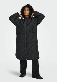 Juodas ilgas pamušaluotas paltas su apvaliu raštu, priekyje su sagomis ir dviem šoninėmis kišenėmis, dėvimas ant juodų kelnių ir batų.