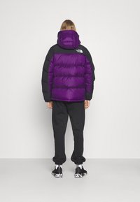 Paarse en zwarte donsjas met capuchon, voorzien van een doorgestikt ontwerp. Draagt over zwarte joggingbroek en zwarte sneakers met een patroon op de zool.