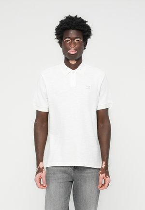 White polo shirt with short sleeves, collared neck, subtle texture, and a small logo patch on the chest; paired with grey jeans.
