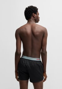 Sorte shorts med en lys blå elastisk livrem med rød "HUGO" tekst. Modellen ses fra ryggen, hvilket fremhæver designet på livremmen.