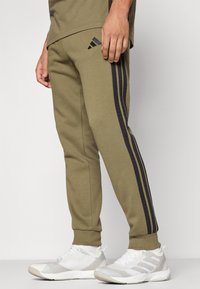 Olivegrønne joggebukser laget av mykt stoff, med svarte sidestrekker, elastisk liv, og ribbede ankler. Kombinert med hvite sportsko.