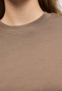 Εγγύς πλάνο ενός ατόμου που φοράει απλό καφέ μπλουζάκι με στρογγυλή λαιμόκοψη, εμφανίζοντας τον λαιμό και το ανώτερο μέρος του στήθους, με μακριά μαλλιά ορατά.