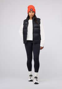 Svart puffervest over en hvit strikkegenser, kombinert med svarte leggings og hvite sneakers. Lyseorange lue, atletisk design.