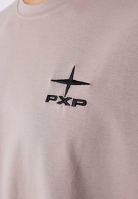 Camiseta de algodón color beige claro con cuello redondo. Presenta un logotipo bordado en negro con una estrella y "PXP" en la parte izquierda del pecho.