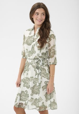 Kobieta w białej sukience do kolan z zielonymi wzorami paisley, z rękawami do łokcia i związanym paskiem w talii, uśmiechająca się i patrząca na bok.
