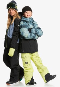 Två barn har på sig vinter sportkläder. Den ena har en jacka med blå och svart mönster tillsammans med svarta byxor; den andra har en jacka i svart och grått och neongula byxor.