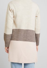 Lång kofta i kräm, brunt och ljusrosa. Gjord av mjukt stickat material med ribbade muddar och splittade sidor för bättre rörelsefrihet.