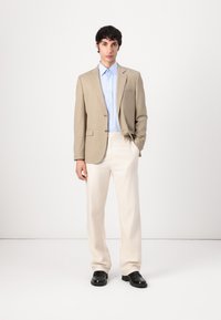 Beige blazer met twee knopen over een lichtblauw shirt, gecombineerd met crèmekleurige broeken en zwarte schoenen. Gladde stof en op maat gemaakte pasvorm.