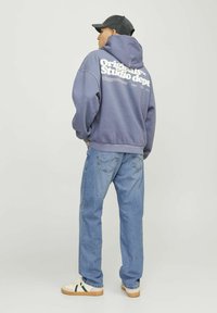 Grauer Kapuzenpullover mit weißem Text auf der Rückseite, kombiniert mit hellblauen Jeans und weißen Sneakers mit grünen Akzenten. Minimalistisches Design.
