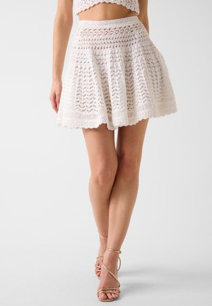 Mini-jupe évasée blanche à motif crochet portée avec des sandales nude à talons hauts et brides, crop top pas entièrement visible.