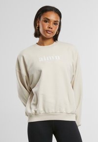 Cremfarbener Pullover mit lockerer Passform, Rippkragen und -bündchen, mit einem weißen Logo auf der Vorderseite. Aus weichem Baumwollmischgewebe.