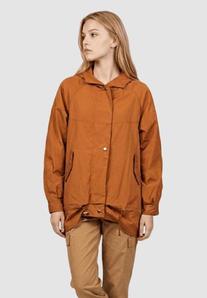 Joven con cabello rubio largo que lleva una chaqueta holgada color naranja quemado y pantalones cargo beige, mirando hacia su izquierda sobre un fondo liso.