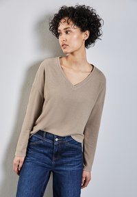 Maglietta beige a maniche lunghe e scollo a V realizzata in tessuto morbido, abbinata a jeans blu scuro con tasche tradizionali e dettagli di cucitura.