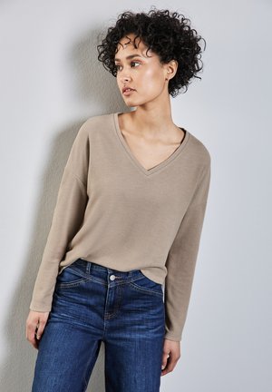 Beige V-Ausschnitt Langarmshirt aus weichem Stoff, kombiniert mit dunkelblauen Jeans mit klassischen Taschen und Nähdetails.