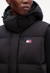 Čierny vypchatý vest s vysokým golierom, na ľavej hrudi má logo Tommy Jeans. Materiály sa zdajú byť jemné s hladkou textúrou.