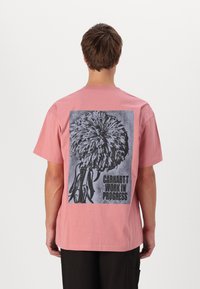 Camiseta de algodón rosa con mangas cortas, que presenta un gráfico de flor negra y el texto "Carhartt Work in Progress" en la parte posterior.