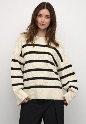 Cremefarbener Strickpullover mit dicken schwarzen Streifen, lässiger Schnitt, gerippte Bündchen und strukturierter Stoff. Das Modell trägt schwarze Hosen.