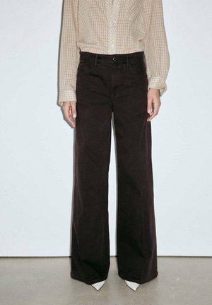 Persona con pantalones negros de talle alto y pierna ancha, camisa beige a cuadros de manga larga metida dentro, y zapatos blancos puntiagudos, de pie contra una pared lisa.