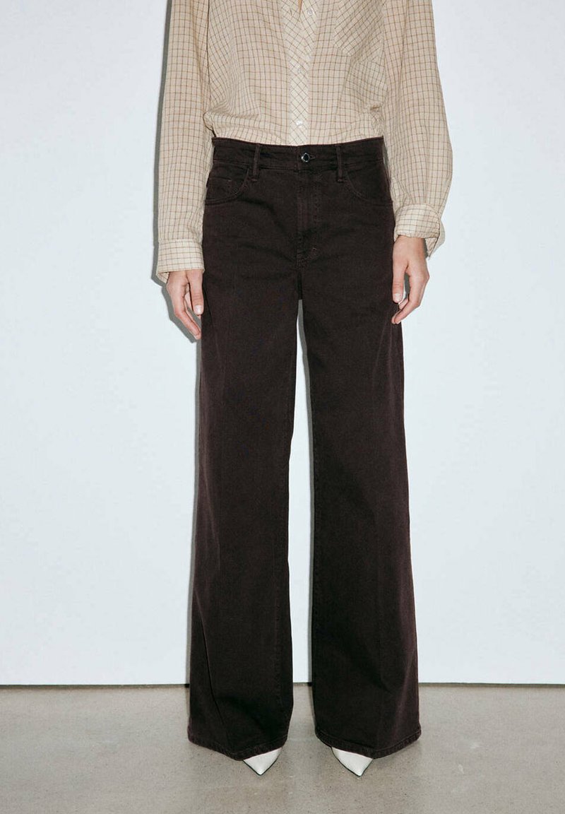 Personne portant un pantalon noir taille haute à jambes larges, une chemise beige à carreaux à manches longues rentrée, et des chaussures blanches pointues, debout contre un mur uni.