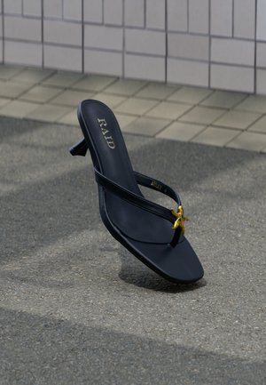 Sandalia negra de tacón con tiras finas y adorno de estrella dorada sobre suelo de asfalto cerca de una pared con azulejos.