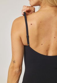 Czarny bezrękawnik z regulowanym paskiem, gładką fakturą i subtelnym połyskiem; widok od tyłu ukazuje odcienie skóry i małe pieprzyki.