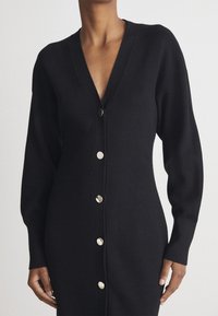 Robe en cardigan en tricot noir avec col en V, manches longues et boutons argentés sur le devant. Texture douce et coupe décontractée.