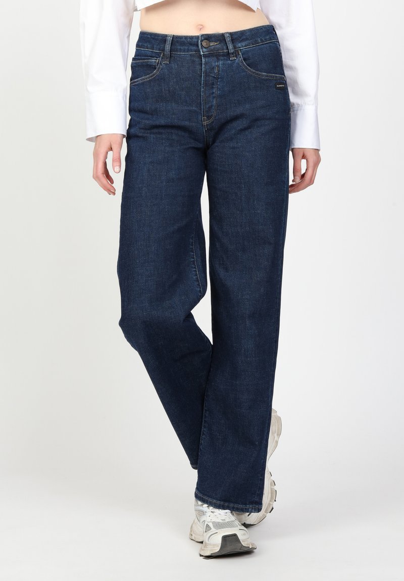 Dunkelblaue Schlagjeans mit hoher Taille, Fronttaschen und kontrastierendem Nähen, kombiniert mit weißen Sneakers. Glatte Denim-Oberfläche.