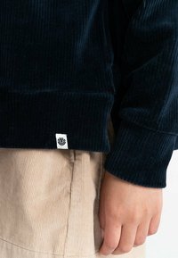Dark blue corduroy sweater with ribbed texture. Features a visible brand label on the hem. Paired with light beige corduroy pants.