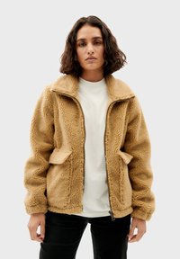 Beige fleece jack met rits aan de voorkant, oversized kraag en twee zakken aan de voorkant. Getextureerd oppervlak; ontspannen pasvorm, gecombineerd met een wit shirt eronder.