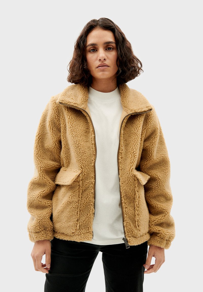 Beige fleece jack met rits aan de voorkant, oversized kraag en twee zakken aan de voorkant. Getextureerd oppervlak; ontspannen pasvorm, gecombineerd met een wit shirt eronder.