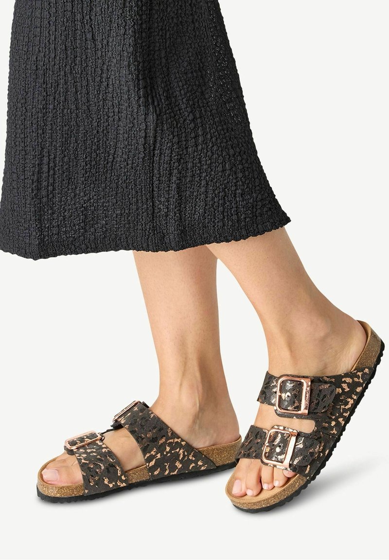 Pieds chaussés de sandales à double bride noires et dorées avec boucles et semelles en liège, assorties à une jupe noire texturée.
