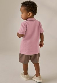 Polo shirt texturizzato rosa chiaro a maniche corte, abbinato a pantaloni corti marroni e scarpe chiare, visto da dietro.